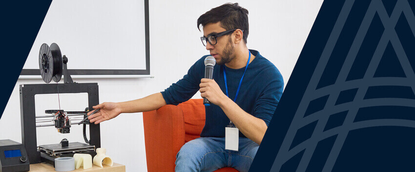 man speaking into microphone pointing at a 3D printer