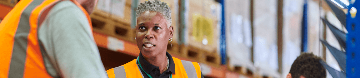 In a warehouse, a woman wearing an orange safety vest over a dark blue shirt stands in the center holding a binder. She is speaking to man on her right, also wearing an orange safety vest. In the background another worker enters data onto a scanner.