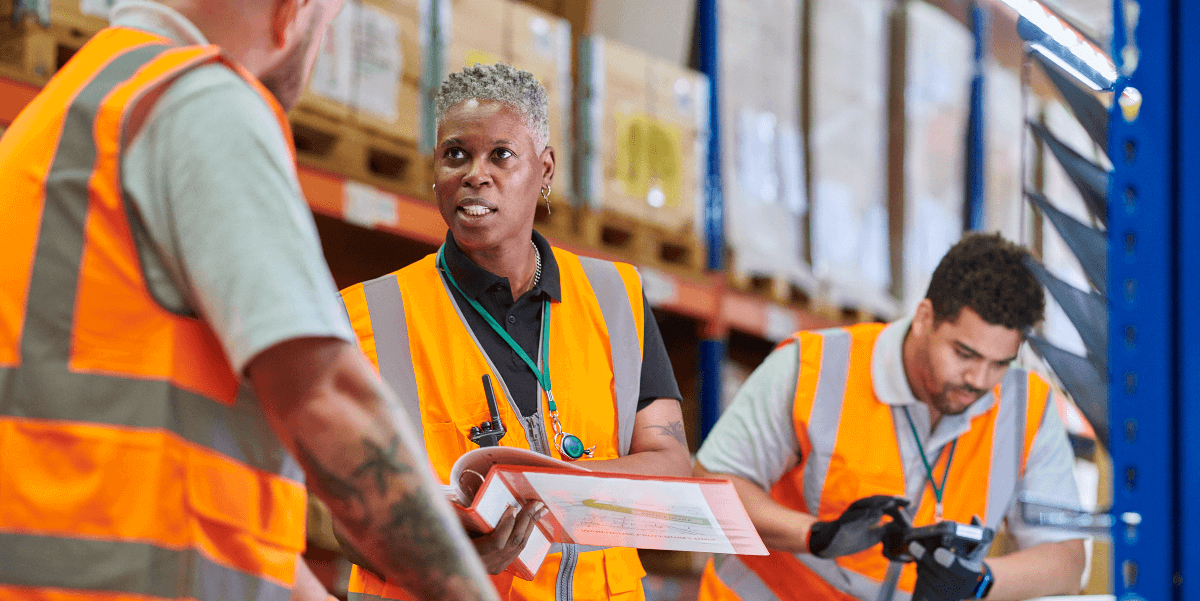 In a warehouse, a woman wearing an orange safety vest over a dark blue shirt stands in the center holding a binder. She is speaking to man on her right, also wearing an orange safety vest. In the background another worker enters data onto a scanner. 