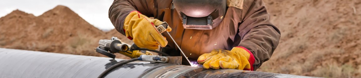 Outdoors with hills of dirt and earth in the background, a pipe fitter wearing wears a heavy brown jacket, welding helmet and orange work gloves, as he welds the joints of a pipe.