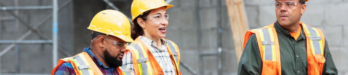 Three construction workers are present in front of a large concrete structure with visible scaffolding, indicating an active construction site. They are all wearing safety gear appropriate for the setting: bright orange vests and yellow helmets. The worker on the left is equipped with a walkie-talkie, likely used for communication on-site. The middle worker is holding an object that is not clearly identifiable from the image. The worker on the right, identified as male, is holding a pair of gloves.