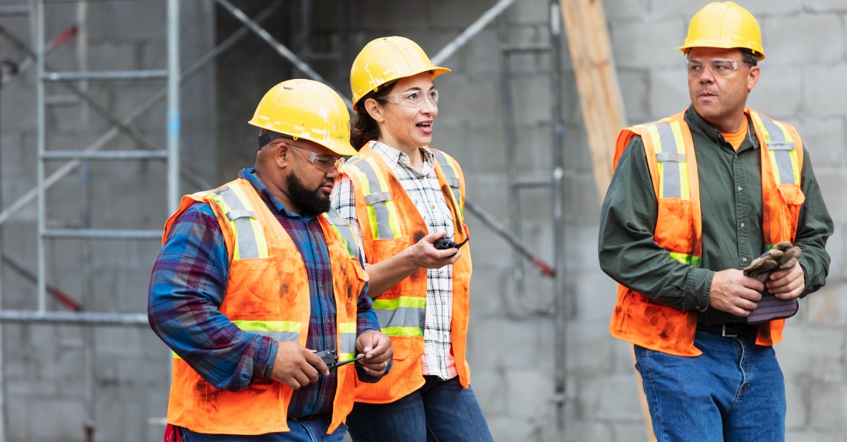 Three construction workers are present in front of a large concrete structure with visible scaffolding, indicating an active construction site. They are all wearing safety gear appropriate for the setting: bright orange vests and yellow helmets. The worker on the left is equipped with a walkie-talkie, likely used for communication on-site. The middle worker is holding an object that is not clearly identifiable from the image. The worker on the right is holding a pair of gloves. 