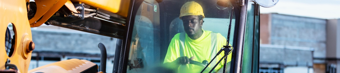 A construction worker operating a yellow excavator on a construction site during twilight hours. The worker is wearing a high-visibility yellow vest and a hard hat, and is seated inside the cab of the excavator, holding the control stick. The background features an industrial setting with buildings under construction and a clear sky at dusk.