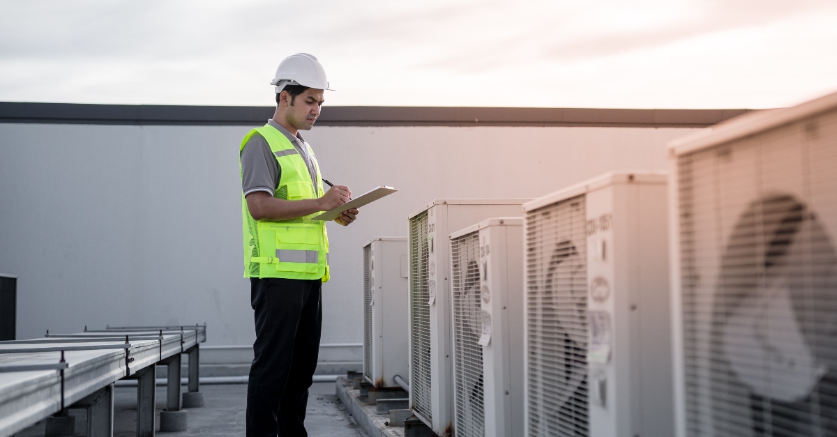 A professional in safety gear, including a helmet and reflective vest, is inspecting or recording details on a clipboard amidst a row of outdoor HVAC units during sunset. The sky is painted with the soft hues of sunset, adding a serene backdrop to the diligent work being performed.