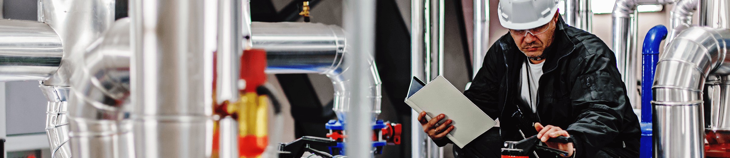 A person inspects stainless steel equipment, holding a clipboard. They are dressed in a black jacket, and the industrial setting is filled with tanks and pipes featuring red and blue valves, indicative of an industrial setting.