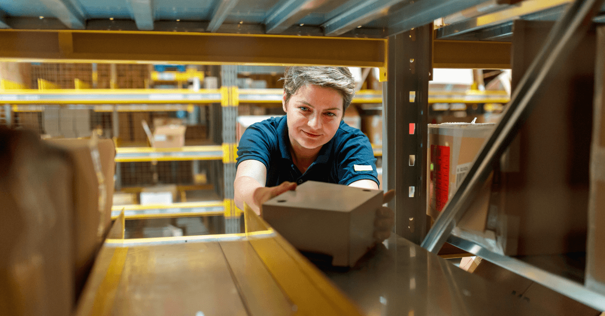 In a warehouse, a woman facing the camera wears a navy shirt and places a box on a nearly full metal shelf.