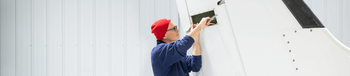 In a well-lit factory, an aviation maintenance technician — wearing a red had, glasses and dark coveralls — stands on a ladder while repairing the tail of an airplane.