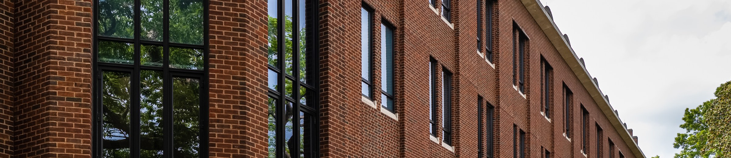 A panoramic view of a red brick building with large, black-framed windows reflecting the surrounding green trees, under a clear blue sky.