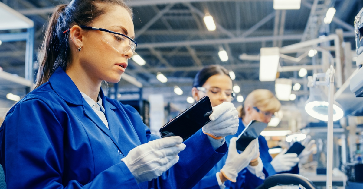 Manufacturing assembly worker wearing a blue work coat and protective glasses inspects a smartphone screen for quality assurance. They are seated at a workstation in a high-tech manufacturing facility with other employees working in the background.