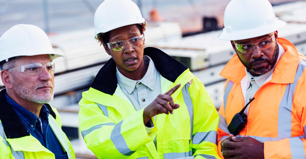 A confident construction foreman stands at a construction site wearing a white hard hat, safety goggles and a reflective vest. They hold a digital tablet and appears to be overseeing the site, suggesting their role as a supervisor or foreman. The background includes construction equipment and materials, emphasizing an active work environment.