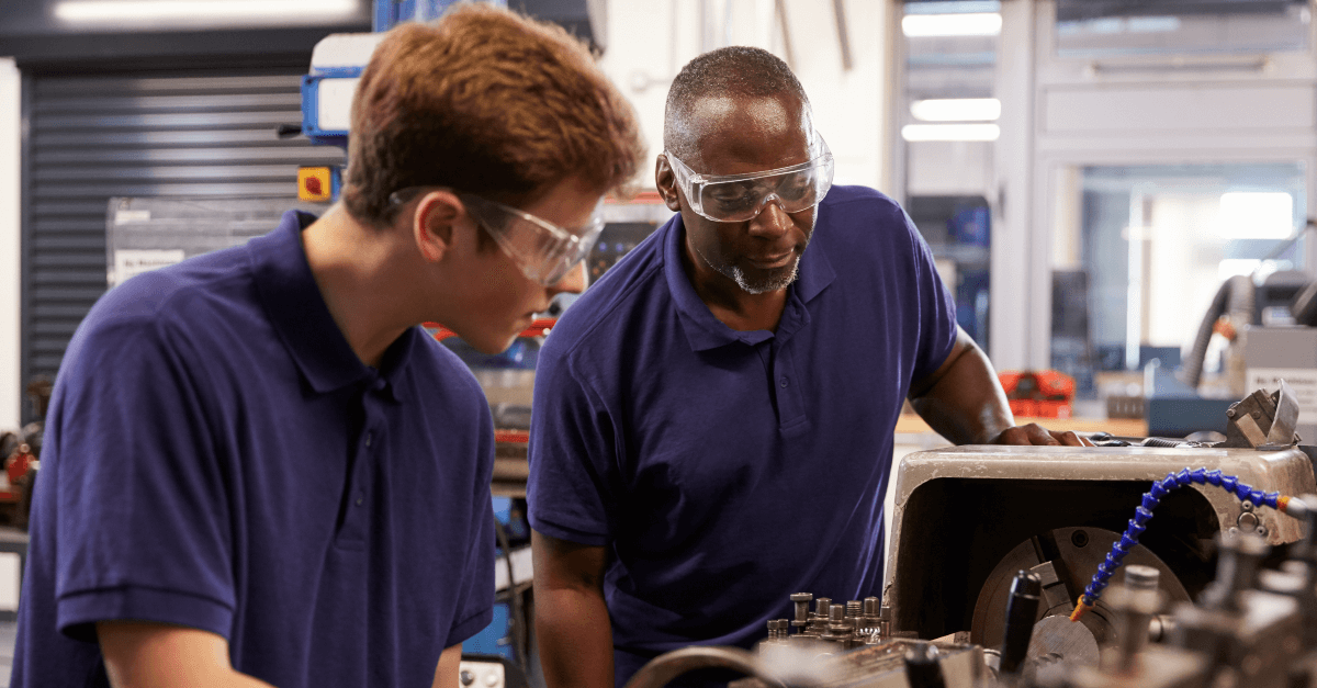 An experienced skilled trades instructor mentors a teenage apprentice on the use of a lathe machine as part of a machinist training program. The setting is an industrial workshop where the instructor, with years of expertise, guides the young trainee through the precise and skilled operation of the machinery. The apprentice, eager to learn, attentively observes the process, gaining hands-on experience that will be invaluable in their future career in advanced manufacturing. This image captures the essence of knowledge transfer in the manufacturing industry, highlighting the importance of practical education in preparing the next generation of skilled machinists.