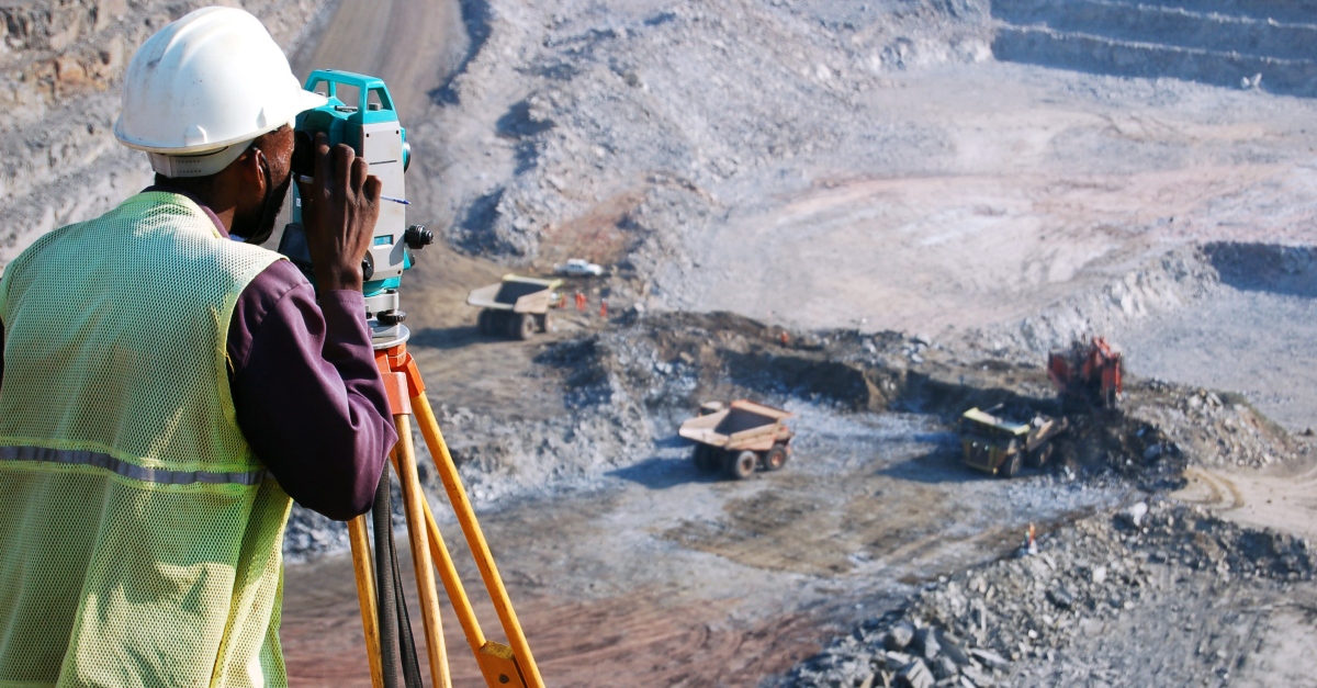 A surveyor in a high-visibility vest and hard hat operates a theodolite at an open-pit mining site. In the expansive background, large dump trucks are actively engaged in the mining operations, highlighting the industry’s massive scale and the meticulous work of surveyors in ensuring operational safety and efficiency.