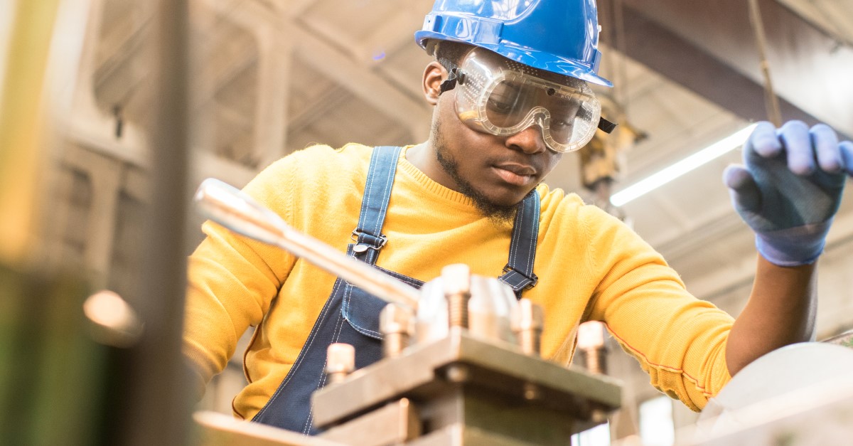 A maintenance worker in overalls and a blue hard hat works on something with his hands