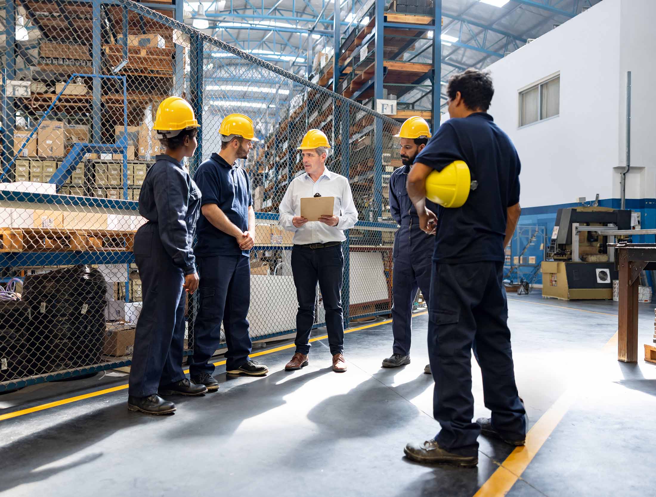 Workers in a food distribution center gather to discuss preventative maintenance. Four workers are wearing dark blue work uniforms and yellow hard hats. One person wears business attire and a yellow hard hat. 