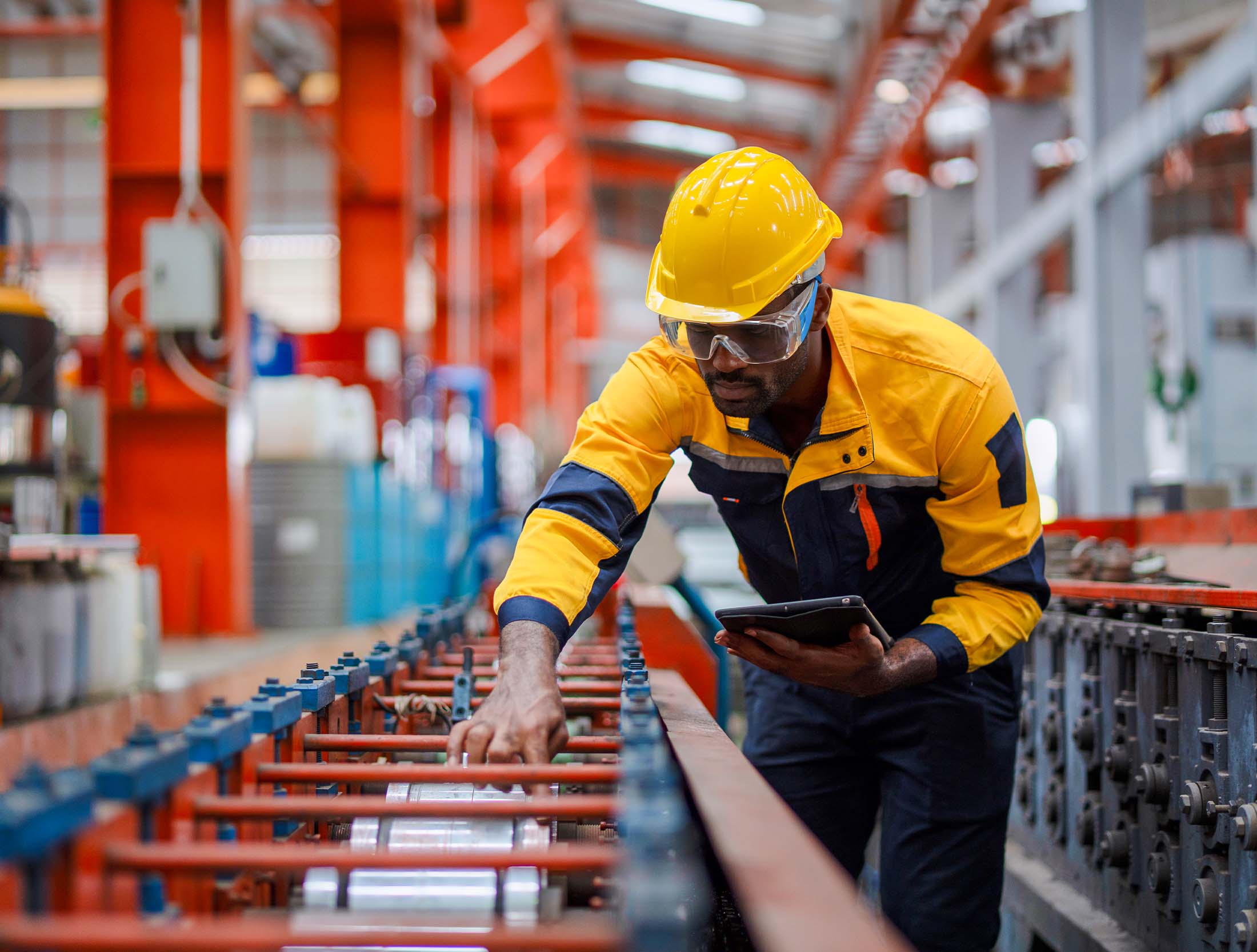 A corrective maintenance worker in a food distribution center inspects a conveyor belt. He wears a yellow and navy safety jacket, safety glasses and a yellow hard hat. 