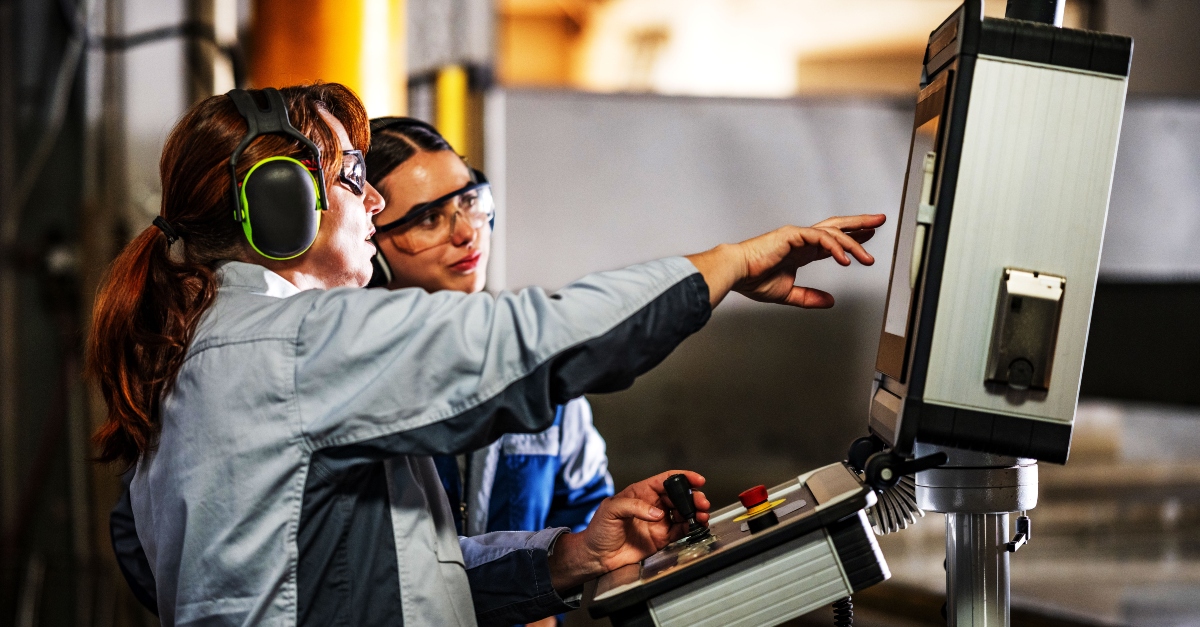 A skilled CNC machinists instructs a younger trainee on operating a CNC waterjet cutting machine in an industrial workshop. Both are wearing safety glasses and work uniforms, focused on the machine's control panel as water sprays in the background.