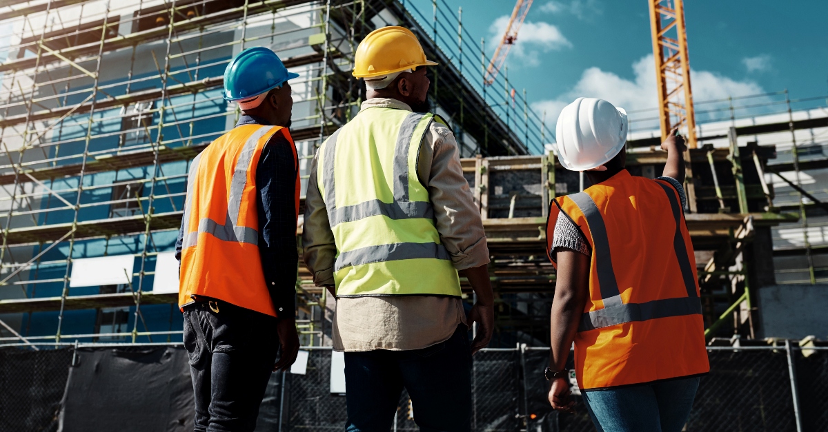 Facing away from the camera, three construction managers wearing orange safety vests and different colored hard hats, look over a jobsite where a crane is lifting objects up towards the scaffolding.
