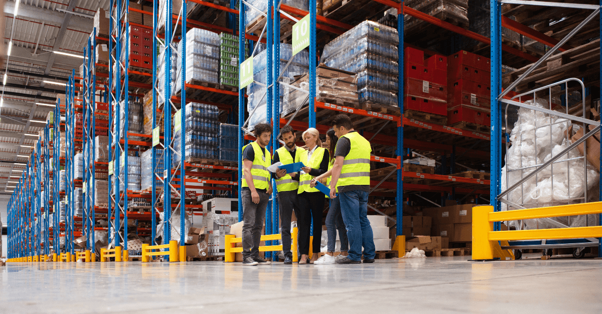 In a warehouse several workers wearing bright yellow safety vests gather around their manager to review a document.
