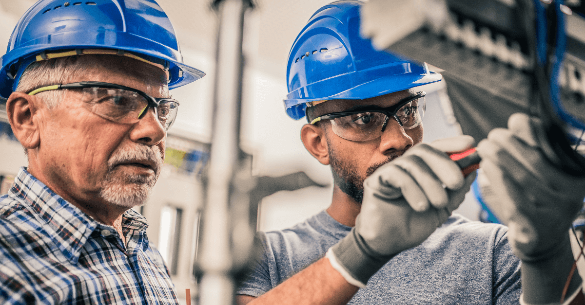 A senior electrician supervises an apprentice connecting the wires in distribution board. Both men are wearing blue hardhats, protective eyewear and safety gloves.