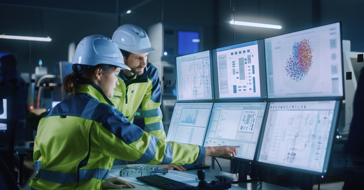 Two maintenance technicians wearing yellow safety jackets and white hard hats operator in a high-tech Industry 4.0 factory, collaborating at a workstation with multiple screens displaying AI-driven production data and machine learning-enhanced assembly processes.