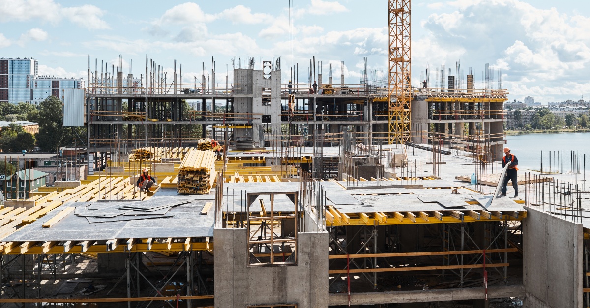 A bustling construction site with multiple cranes and workers. The scene includes various construction equipment and materials, with workers wearing safety gear such as helmets and vests. The background shows partially constructed buildings and scaffolding, indicating ongoing development. Contact Aerotek to learn more about employee performance monitoring solutions.