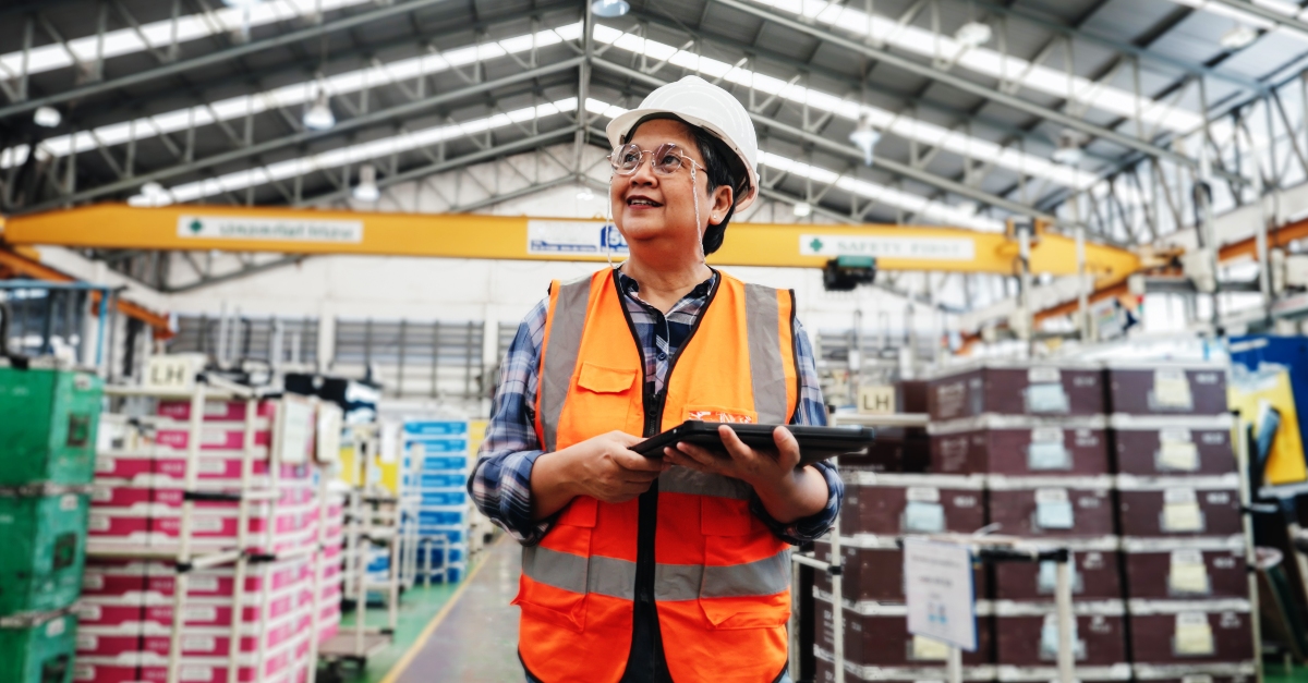 A facilities technician wearing a white hard hat and yellow safety vest, holds a tablet while touring a distribution facility. 