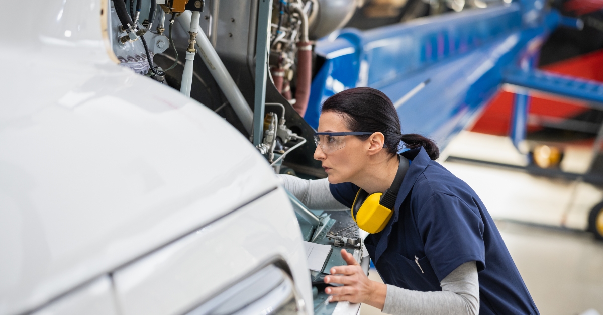 Female aircraft mechanic with a digital tablet, meticulously inspecting and working on a helicopter inside a hangar. The setting conveys a theme of safety and precision in aviation maintenance