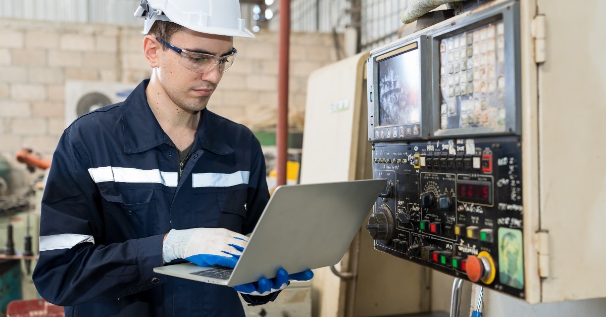 A maintenance technician in a high-visibility vest and hard hat working on a laptop computer within an industrial environment, with various tools and equipment in the background.