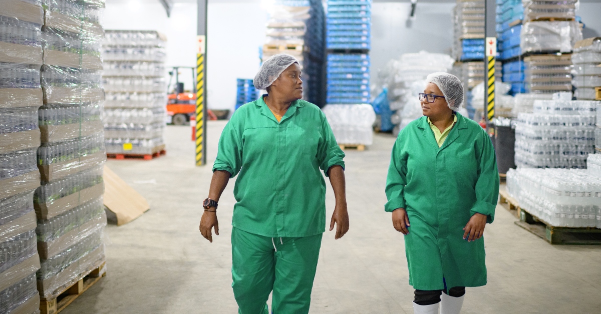 Two women in protective workwear inspecting water bottles on a production line inside a bottling warehouse as part of a quality control process. This image displays jobs in the food and beverage industry. 