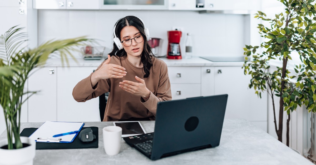 A woman sitting at a kitchen table is conducting a video job interview. She wears white headphones as she uses her hands to emphasize hands-on experience. 
