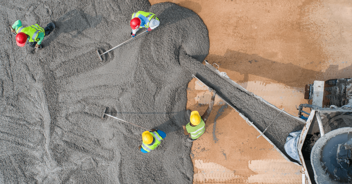 An aerial shot shows a concrete mixer pouring concrete onto dirt as four workers wearing hard hats and safety vests use tools to smooth the concrete before it sets. 