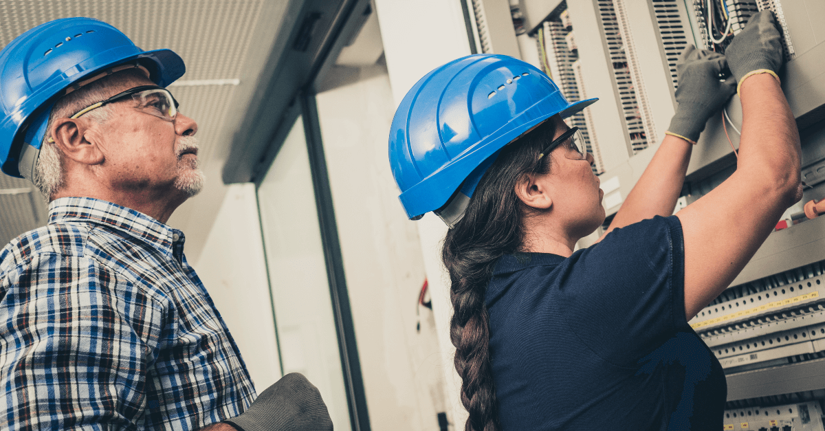 In a small room, a man and a woman wearing blue hard hats work on an electrical box on a wall.