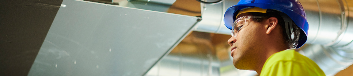 In a factory, a man wearing a blue hard hat, safety glasses, yellow reflective shirt and gloves looks at a tablet while inspecting industrial ductwork.