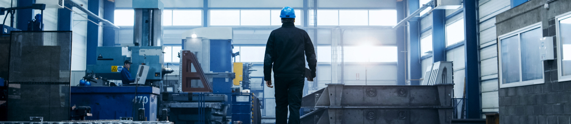 An industrial setting with a person in a hard hat walking toward a bright entrance. The spacious interior features high ceilings, machinery, and an overhead crane system. Cool blue tones suggest early morning or late afternoon