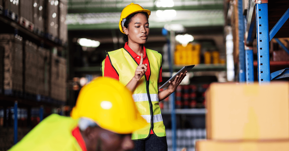 In a warehouse a man and woman wear hard hats and safety vests while logging inventory data into tablets.