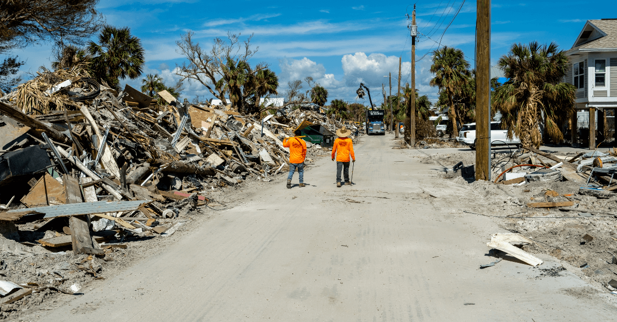On a sunny day, two workers walk away from down a dirt covered road facing away from the camera. They are wearing orange long-sleeve shirts, blue denim pants and work boots. To their left, piles of debris line the street. To their right, several homes and cars remain intact. Aerotek can help companies overcome staffing challenges in disaster affected areas.