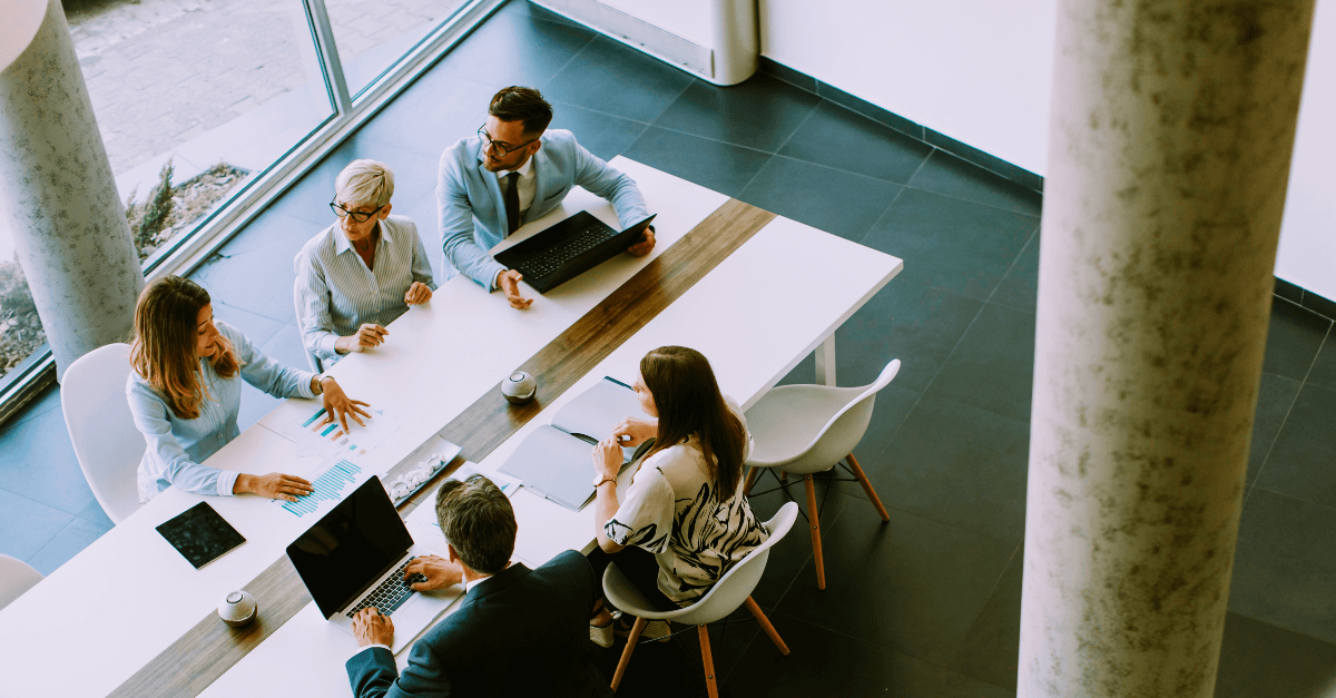 An overhead view of a business meeting shows 5 people having a discussion at a long white table.