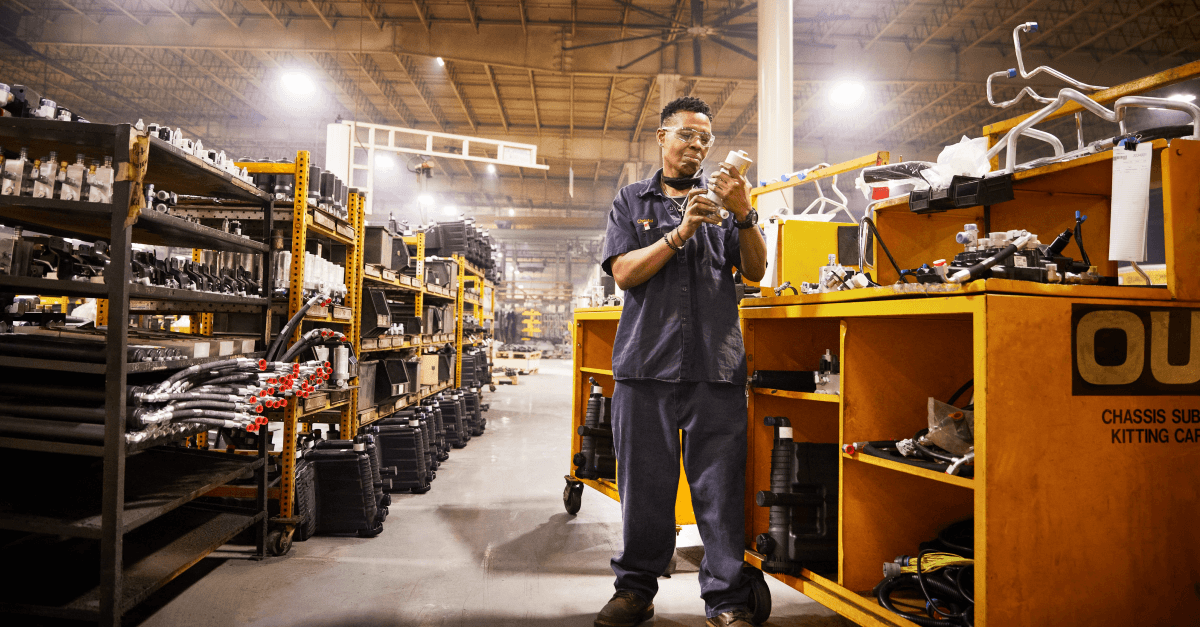 A woman wearing dark work clothes and safety glasses closely inspects a replacement component. 