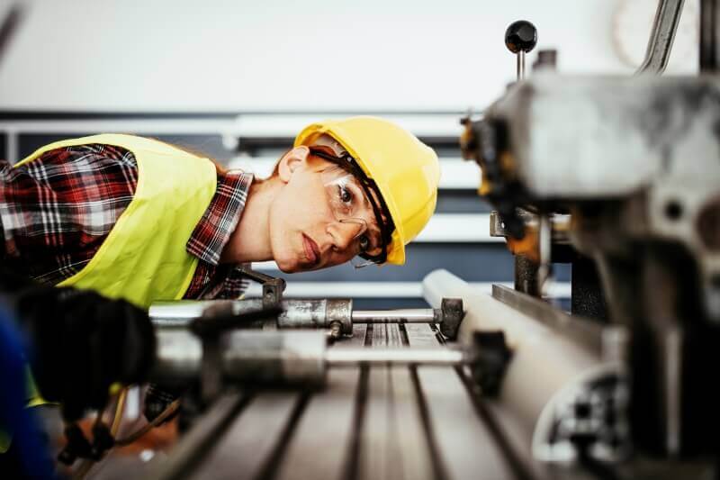 A woman wearing protective earwear, hardhat, safety vest and red flannel shirt inspects drilling machine.