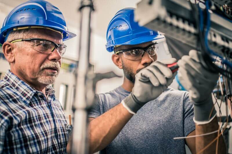 A senior electrician supervises an apprentice connecting the wires in distribution board. Both men are wearing blue hardhats, protective eyewear and safety gloves.