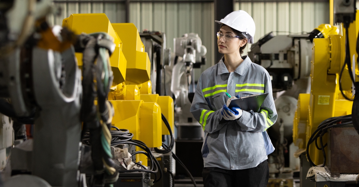 A maintenance technician wearing a safety helmet and high-visibility vest for safety, is inspecting a machine in a factory setting. She is focused on checking the equipment, ensuring everything is functioning correctly to ensure safe and efficient operations. 