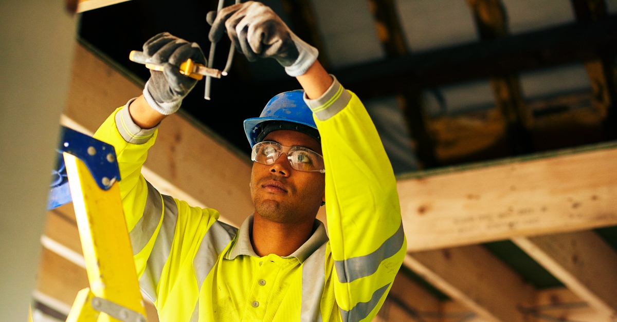 A young master electrician wearing a gray hoodie and safety glasses, working attentively on electrical wiring inside a partially renovated space. He is using tools to adjust wires within an open wall cavity, surrounded by exposed wooden framing and insulation.