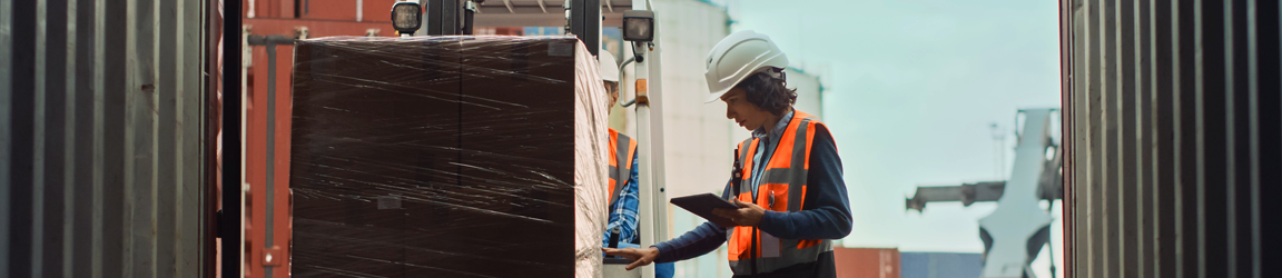 A warehouse worker wearing a white construction hat and orange vest examines a pallet on a job site