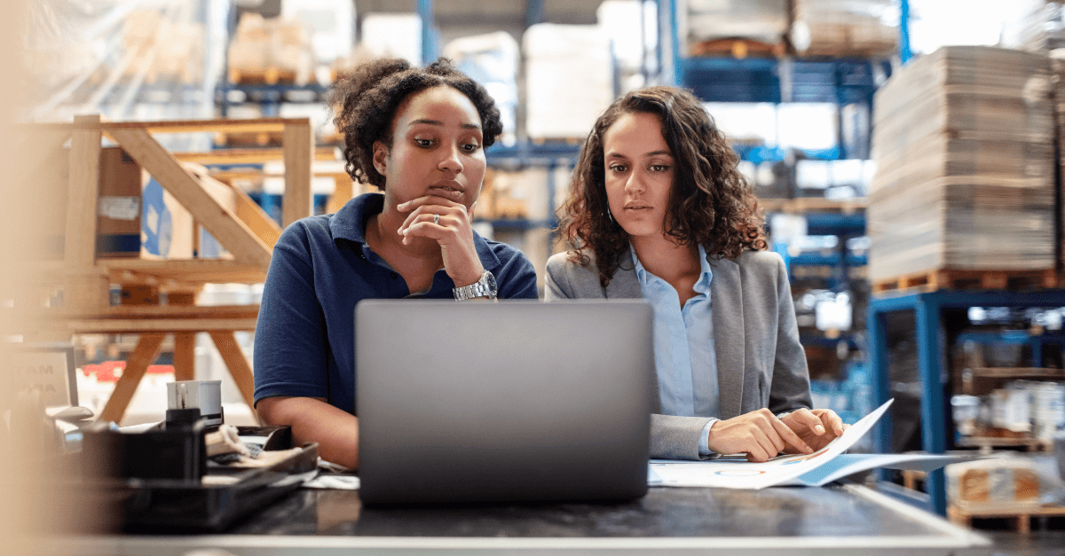Two female workers in a logistics environment look at a laptop