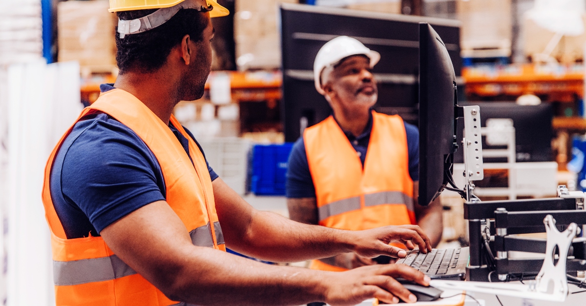 Inside a warehouse, two picker packers (or warehouse associates) in safety vests and work attire review daily duties among storage racks and packaged goods.