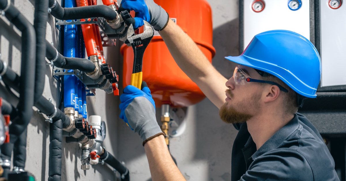 A plumber repairs a plumbing system, using a wrench to adjust metal pipes connected to a wall-mounted unit. He is wearing a gray shirt and safety gloves, working in a well-lit, unfinished room with exposed walls.