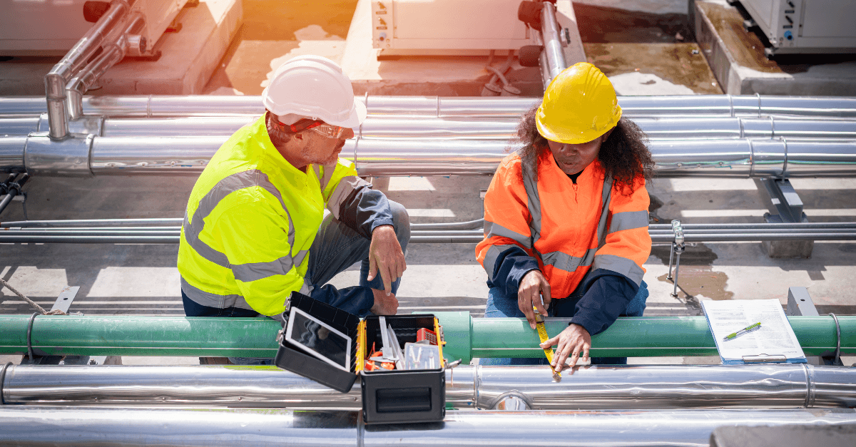On a roof, a man and woman wear yellow safety jackets while kneeling over several metallic pipes. On the left, the man wears a white hardhat and faces the kneeling woman wearing an orange hardhat on the right. Aerotek can help you find the right HVAC technician job.