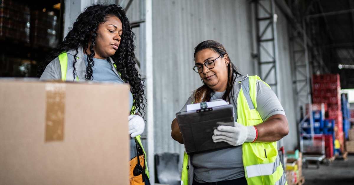 Two warehouse workers in safety vests checking and scanning cardboard boxes on metal shelving racks in a well-lit industrial distribution facility.