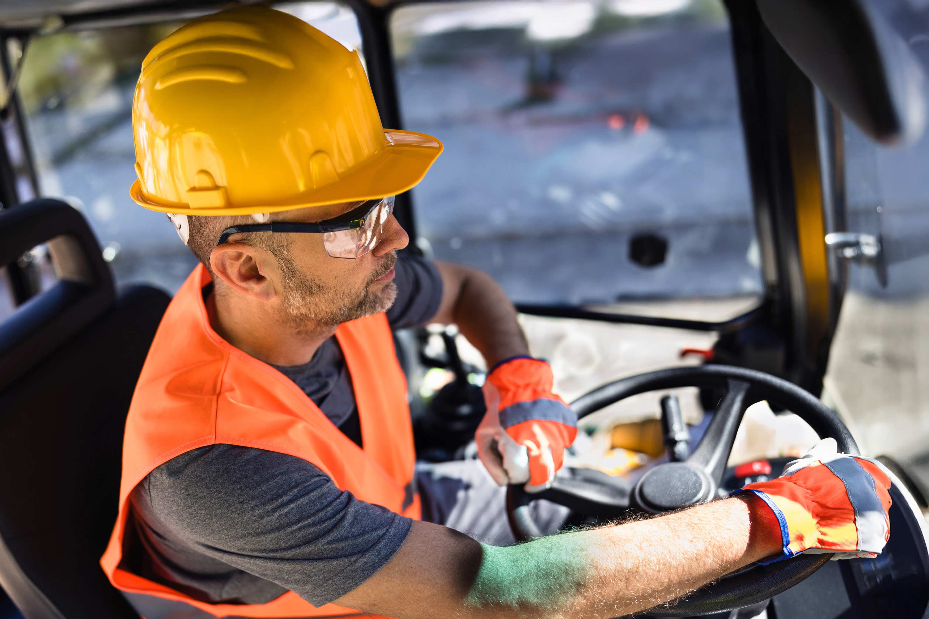 A heavy equipment operators turns the steering wheel while wearing safety glasses, vest, gloves and hard hat as he works on a data center construction site. 