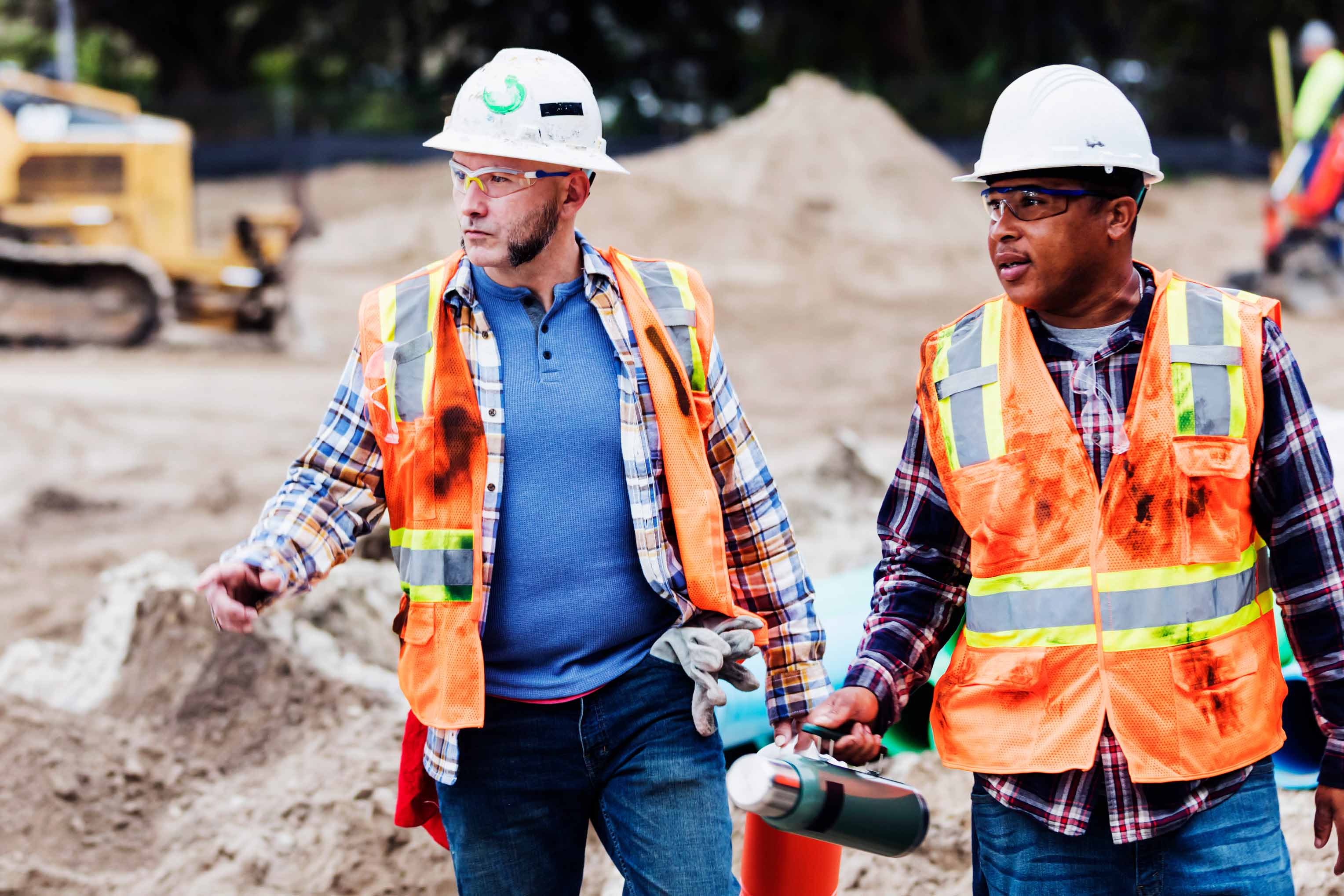 Construction professionals wearing hard hats, safety glasses and high-visibility vests walk through an active jobsite with heavy equipment and earthmoving work in the background.